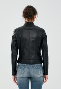Zwarte leren jas met een getailleerd ontwerp, schoudervulling en zichtbare stikseldetails. Draagt over blauwe jeans met een kenmerkend zakpatroon.