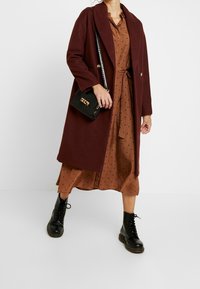 Burgunderroter Wollmantel über einem braun gepunkteten Kleid mit tailliertem Schnitt. Eine schwarze Umhängetasche und schwarze Schnürstiefel runden den Look ab.