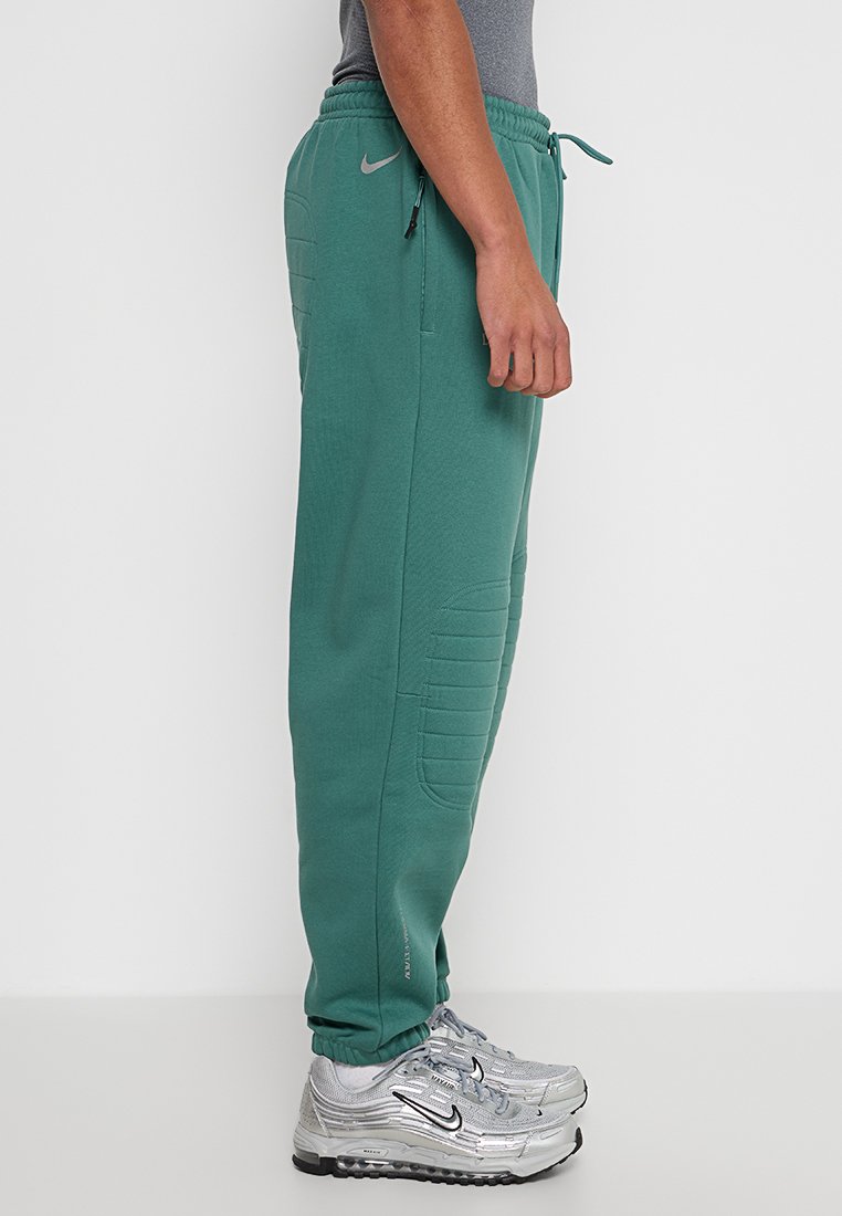 Persona che indossa pantaloni felpati Nike color turchese e scarpe Nike argentate, in piedi di lato contro uno sfondo bianco semplice.