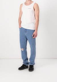 Hombre de pie con las manos en los bolsillos, vistiendo una camisa sin mangas blanca, unos pantalones vaqueros rotos de color azul claro y zapatillas negras.