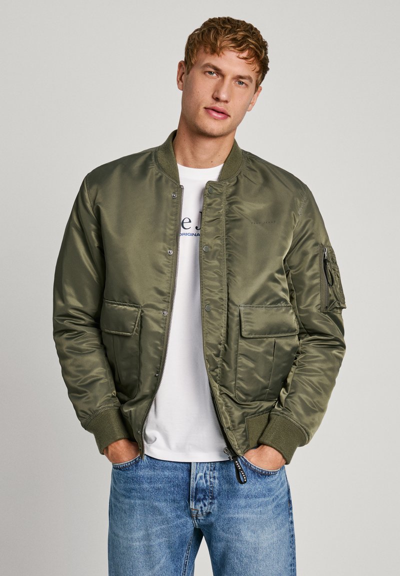 Chaqueta bomber de color verde oliva, hecha de una tela suave y brillante. Cuenta con dos bolsillos frontales, un bolsillo con cremallera en la manga y puños acanalados.