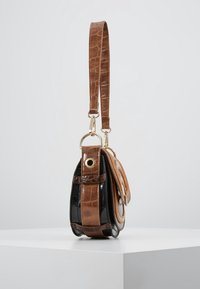 Mała czarna i brązowa torebka skórzana w wzór krokodyla z złotymi akcentami i pętelkowym brązowym paskiem, wyeksponowana na białym piedestale.