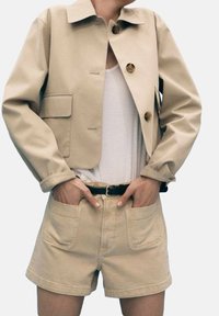 Chaqueta cropped beige con botones redondos, bolsillo frontal, combinada con pantalones cortos beige a juego que tienen dos bolsillos delanteros. Camiseta blanca lisa debajo.