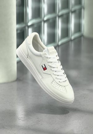 Baskets en cuir blanc avec lacets, bout perforé, et petit logo rouge, blanc et noir sur le côté, flottant au-dessus d'un sol gris.