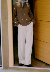 Femme debout dans un encadrement de porte portant un pull marron à pois, une écharpe grise, un pantalon blanc à jambes larges et des chaussures noires, les mains dans les poches.