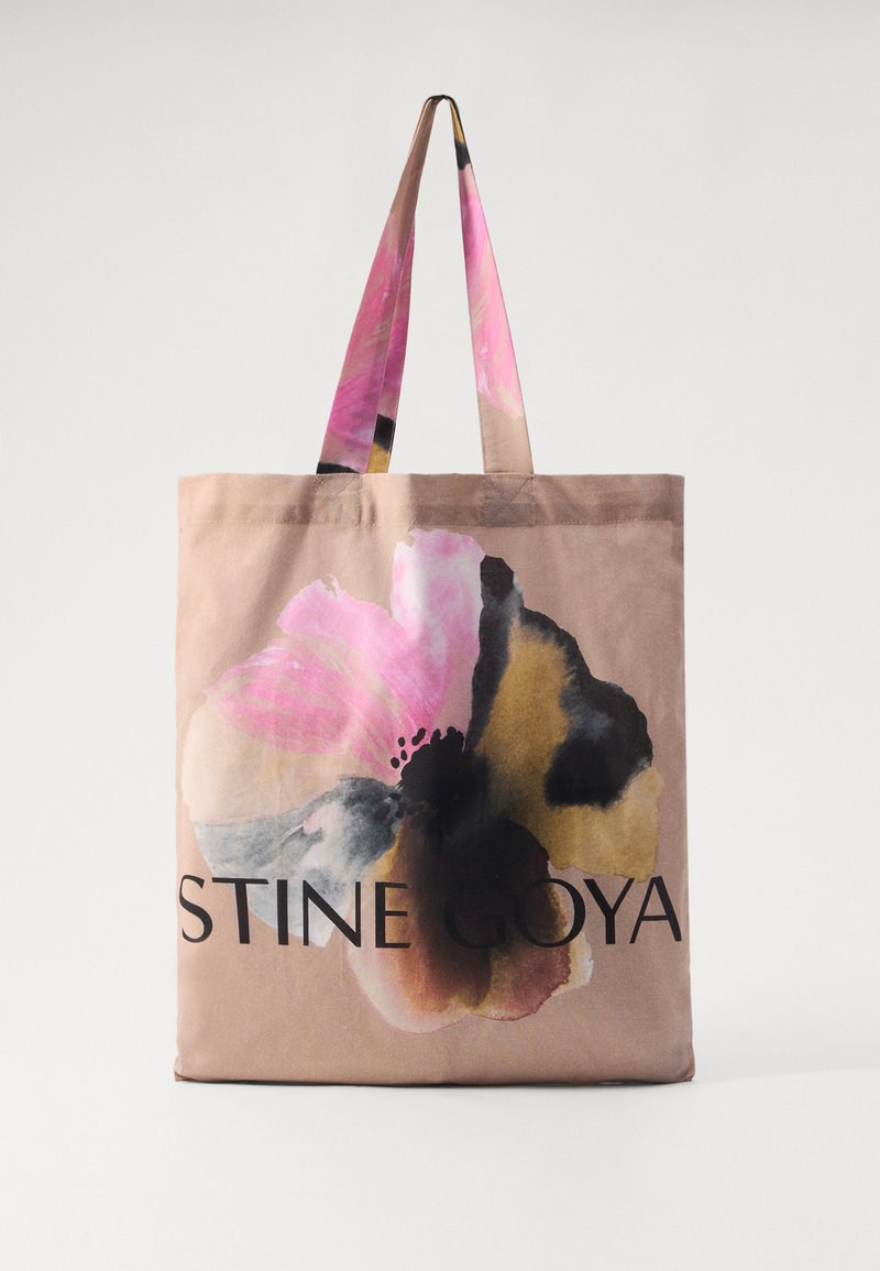 Canvas tote bag in beige with a large pink and black floral print. Features long handles and the brand name "STINE GOYA" in bold black text.