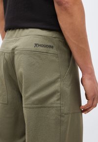 Κοντινό πλάνο ενός ατόμου που φοράει χακί σορτς με μαύρο κέντημα λογότυπου "HOUDINI" στην πίσω τσέπη και μαύρο μπλουζάκι.