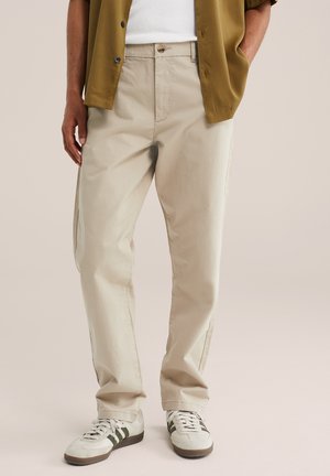 Man met beige rechte broek, witte sneakers met zwarte strepen, wit overhemd en open olijfgroen overhemd, handen in de zakken.