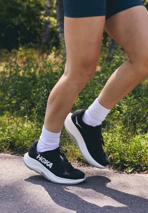 Pantofi de alergare negri cu logo și accente albe, având o talpă groasă, căptușită și un design rotunjit. Purtați cu șosete albe până la glezne.