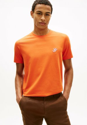 Młody mężczyzna w pomarańczowej koszulce z krótkim rękawem z małym białym logo na piersi i brązowych spodniach, ręce w kieszeniach, neutralne tło.
