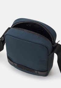 Piccola borsa quadrata in materiale sintetico di colore blu scuro, con chiusura a zip, tracolla regolabile e una fascia nera orizzontale sul davanti.