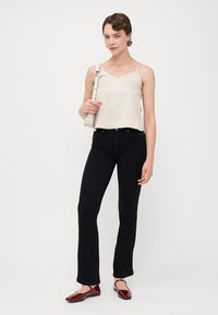 Krämfärgad satинspets, svarta utsvängda jeans och bordeauxfärgade tofflor. Modell som håller en vit handväska, med kort hår och hoopsörhängen.