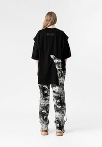 Persona de espaldas vistiendo una camisa negra grande con un diseño abstracto de mano en blanco y negro, y pantalones a juego con un patrón, complementados con zapatos beige.