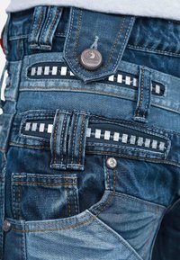 Jeans in denim con lavaggio blu scuro, cuciture visibili in arancione, dettagli decorativi in argento e numerose tasche tra cui una tasca con zip.