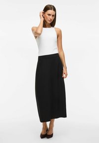 Jupe maxi noire en tissu lisse avec une taille fine, associée à un top sans manches blanc. Le mannequin se tient en talons, main dans les cheveux.