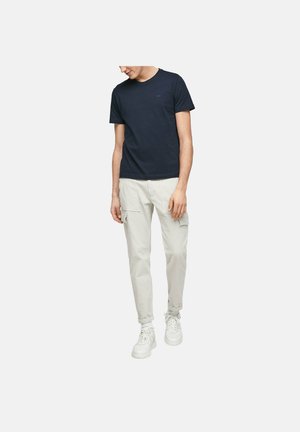 Homme portant un t-shirt bleu marine foncé à manches courtes, un pantalon cargo beige clair, des baskets blanches, debout devant un fond clair uni.