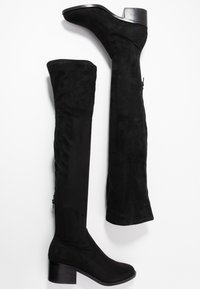 Bottes en suède noir au-dessus du genou avec un talon épais, présentant une texture lisse et un détail de fermeture éclair sur le côté.