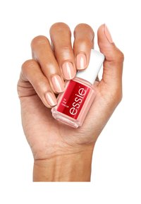 Bottiglia di smalto Essie tenuta in una mano, con una base in vetro quadrata, un tappo bianco e un'etichetta rossa; unghie verniciate di un raffinato smalto rosa.