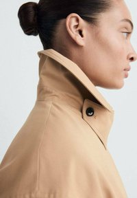 Trench coat bej deschis cu guler înalt, având detalii cu nasturi negri. Textură de țesătură netedă cu o croială ajustată, prezentată din lateral.