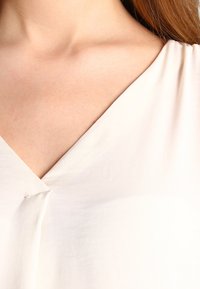 Primo piano del collo e della parte superiore del torace di una persona che indossa un indumento bianco a scollo a V, mostrando il tono della pelle e la texture liscia del tessuto.