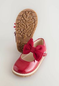 Scarpa in pelle sintetica rossa con un grande fiocco testurizzato, suola beige con motivo a foglia e cinturino alla caviglia con fibbia metallica.