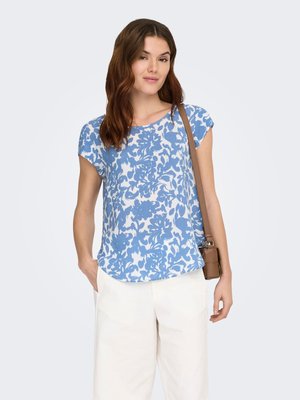 Femme portant un haut à fleurs bleu et blanc à manches courtes, un pantalon blanc et un sac bandoulière marron, debout avec une main dans la poche.