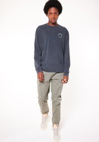 T-paita pitkällä hihalla tummanharmaana, jossa on pyöreä logo vasemmassa rinnassa, yhdistettynä vaaleanvihreisiin cargo-housuihin ja beigeihin korkeisiin tennarikenkiin.