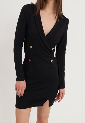 Femme portant une robe style blazer noire à manches longues avec des boutons dorés et une fente avant, debout devant un fond clair uni.