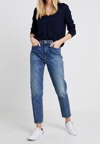 Haut à manches longues bleu foncé associé à un jean taille mi-haute bleu clair. Le mannequin porte des baskets blanches. Tissu lisse, coupe droite.