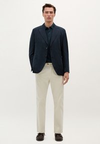 Námořnický lněný blazer s dvěma knoflíky, kombinovaný s tmavým polo tričkem a béžovými chinos kalhotami. Hnědé šněrovací boty dotvářejí celý outfit.