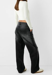 Schwarze weit geschnittene Jeans mit einem verblassten Finish, ausgestattet mit Gesäßtaschen und einer hohen Taille, kombiniert mit einem enganliegenden weißen Langarmoberteil.