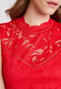 Top sin mangas de encaje rojo que presenta un elegante escote alto con bordes en forma de scallop y patrones florales intrincados, con un acabado suave y texturizado.