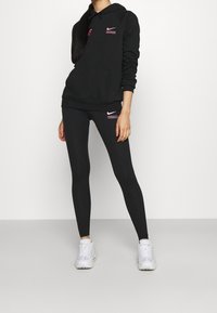 Svart hoodie och leggings, båda med rosa Nike-logotyper. Hoodien har en framficka och leggingsen har en horisontell logodetalj. Vita sneakers bärs.