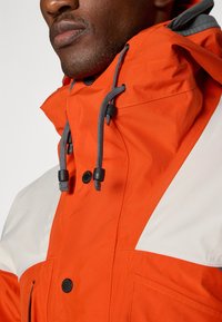 Orange waterproof jacket with gray accents, featuring a high collar, adjustable drawstrings, and button closures. Soft texture and clean design.