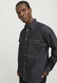 Camicia in denim scuro con chiusura a bottoni, due tasche sul petto e cuciture a contrasto. Presenta un colletto tradizionale e maniche lunghe.
