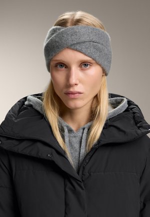 Brățișă gri tricotată cu un design răsucit, purtată cu o jachetă puffer neagră și un hanorac gri. Textură moale, aspect confortabil.