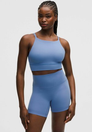 Kobieta z warkoczami, ubrana w dopasowany jasnoniebieski krótki top i rowerowe szorty z wysokim stanem na jednolitym tle.