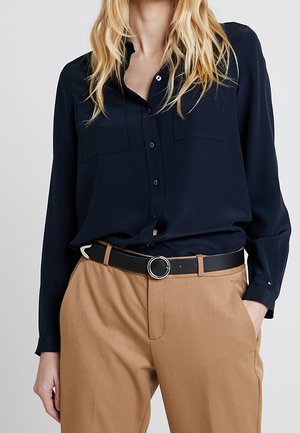 Kvinde iført en mørkeblå skjorte med knapper, stoppet ned i kamelfarvede bukser med et sort bælte med en rund, sølvfarvet spænde.