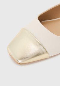 Beige platt sko med en fyrkantig metallisk guld tåhätta. Ovandelen är slät och innersulan är ljusbrun. Har en sömlös design.