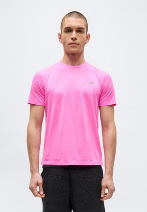 Jeune homme aux cheveux courts portant un t-shirt à manches courtes New Balance rose vif et un short noir texturé, debout devant un fond uni.