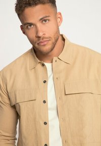 Camisa de botones color tostado hecha de una tela ligera, con dos bolsillos en el pecho, botones negros y un cuello clásico. Usada sobre una camisa blanca.