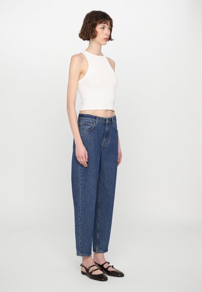 Top corto blanco sin mangas, jeans azul de cintura alta y zapatos planos oscuros. Los jeans tienen cinco bolsillos y un diseño de pierna recta.