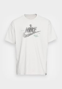 Valkoinen puuvillapaita, jossa on graafinen kuva mustista juurista, jotka on kiedottu sanan "Nike" ympärille, ja teal-värinen teksti "Kasvu urheilussa."