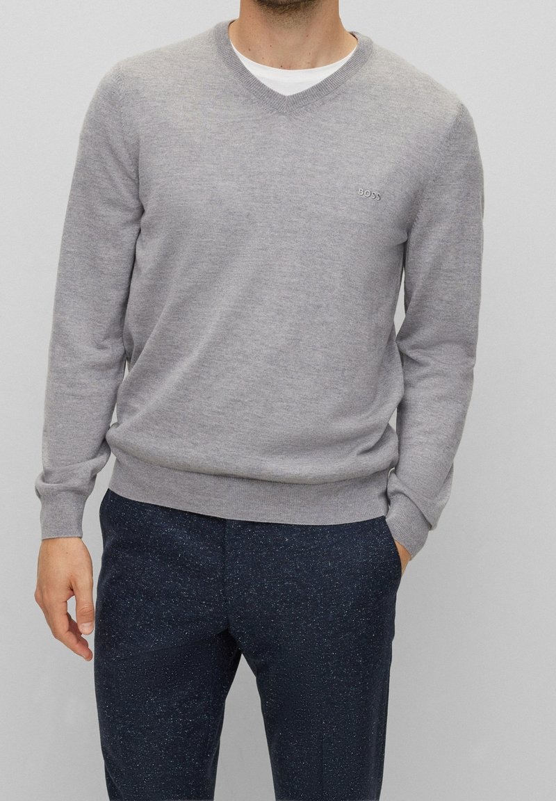 Pull gris en V en tissu doux, avec des poignets et un ourlet côtelés, et un petit logo brodé sur la poitrine. Associé à un pantalon sombre.