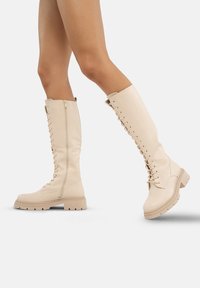 Botas de cuero beige hasta la rodilla con cordones y cremallera lateral, suelas gruesas y parte superior texturizada. Presentan refuerzo alrededor del área de los cordones.