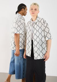 Schwarzes und weißes, gemustertes Kurzarmhemd mit einem geometrischen Spitzenmuster, getragen mit Jeans und einem schwarzen Rock, mit einem Kragenaufschlag.