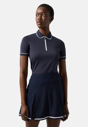 Woman wearing a navy blue short-sleeve polo shirt with white trim and a navy pleated skirt with white accents, standing against a plain background.