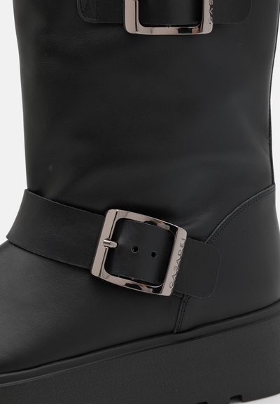 Bottine en cuir noir avec une large sangle et un grand détail en boucle métallique. Présente une forme structurée et une finition texturée.