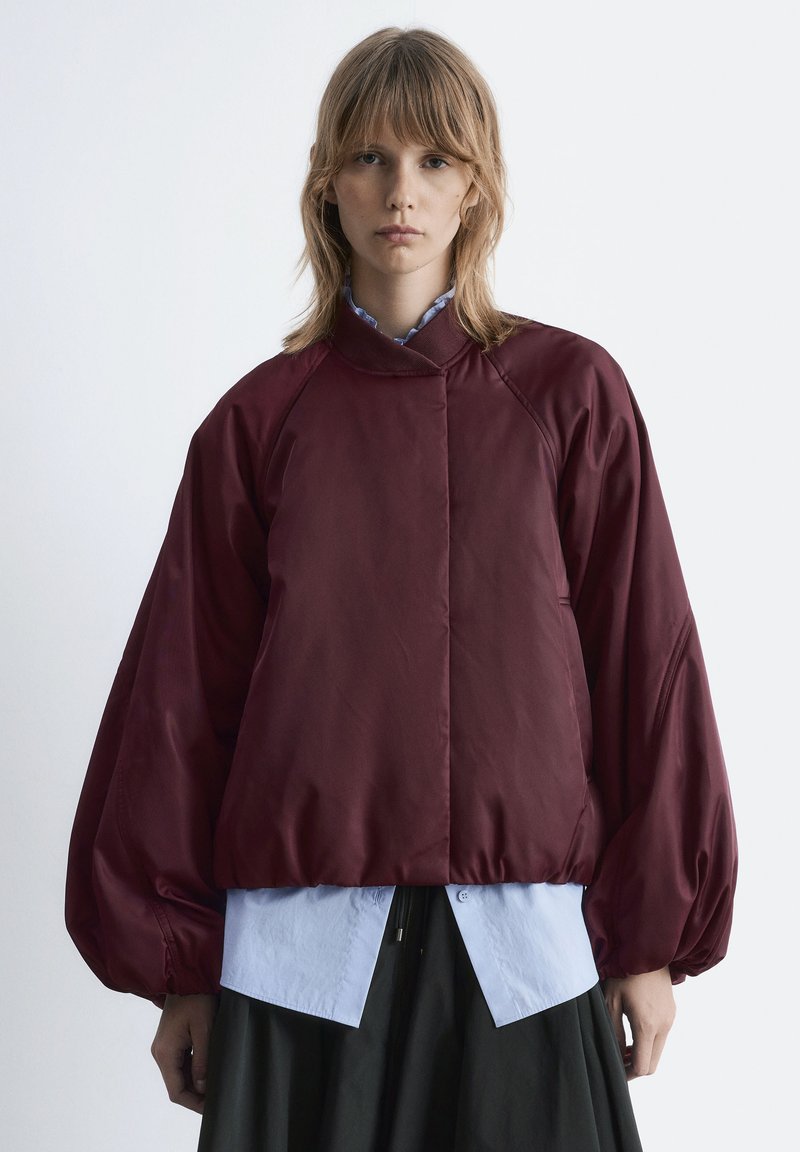 Burgunderfarbene gepolsterte Jacke mit hohem Kragen, Raglanärmeln und elastischen Bündchen. Getragen über einem hellblauen Hemd und einem dunklen Rock mit glatter Textur.