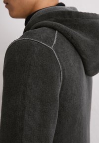 Grijze hoodie met een textuur in de brei, voorzien van een trekkoordcapuchon en contrasterende stiksels langs de naden. Close-up weergave van de zijkant.